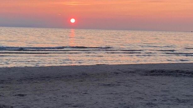 Auringonlasku rannan yllä, jossa vesi on tyyntä ja taivas kirkas.