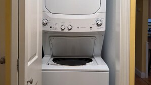 A compact laundry room with a washer and dryer, a door, and a small storage area above the dryer. - Quaint 2-bedroom house overlooking Lake Winnipesaukee in welcoming Gilford (Gilford)