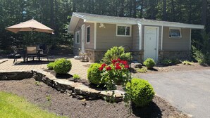 A single-story mobile home with a patio area, outdoor seating, and a stone retaining wall with landscaping. - Quaint 2-bedroom house overlooking Lake Winnipesaukee in welcoming Gilford (Gilford)