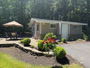A single-story mobile home with a patio area, outdoor seating, and a stone retaining wall with landscaping.