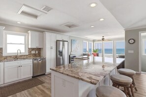 A modern kitchen with white cabinetry, a large island with stools, a stainless steel refrigerator, and a view of the ocean through large windows. - Mayor's Penthouse: Oceanfront luxury w/elevator 4BR (Carolina Beach)