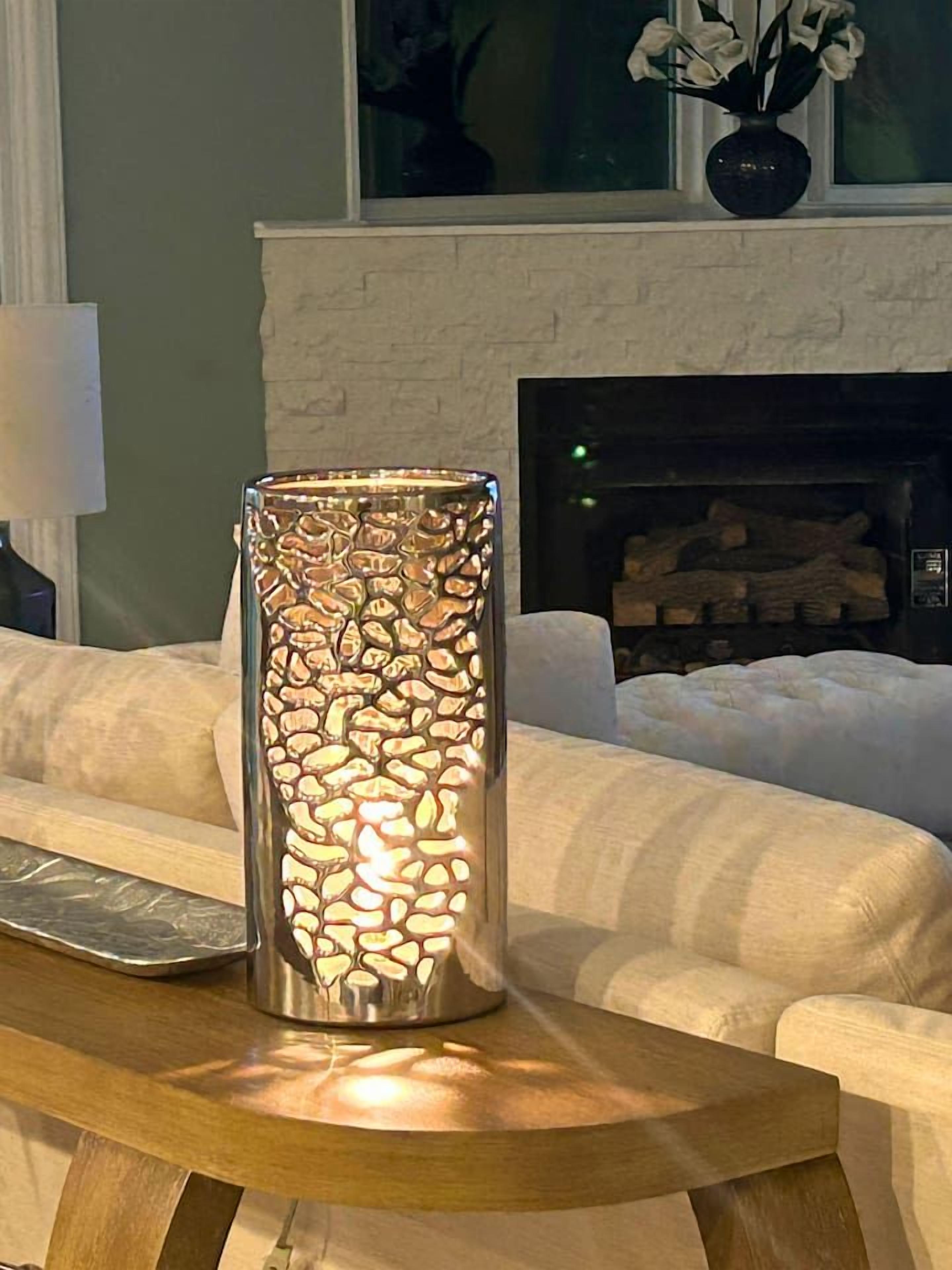 A tall, textured candle holder on a wooden table in a living room with a fireplace in the background.