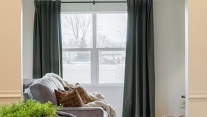 Un espace repas avec une longue table en bois, des chaises noires, un lustre et une vue sur l’extérieur à travers une fenêtre ornée de rideaux.