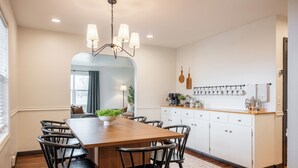Un espace repas comprenant une table en bois, des chaises noires, un lustre, ainsi qu’un coin cuisine avec des meubles blancs et un plan de travail.