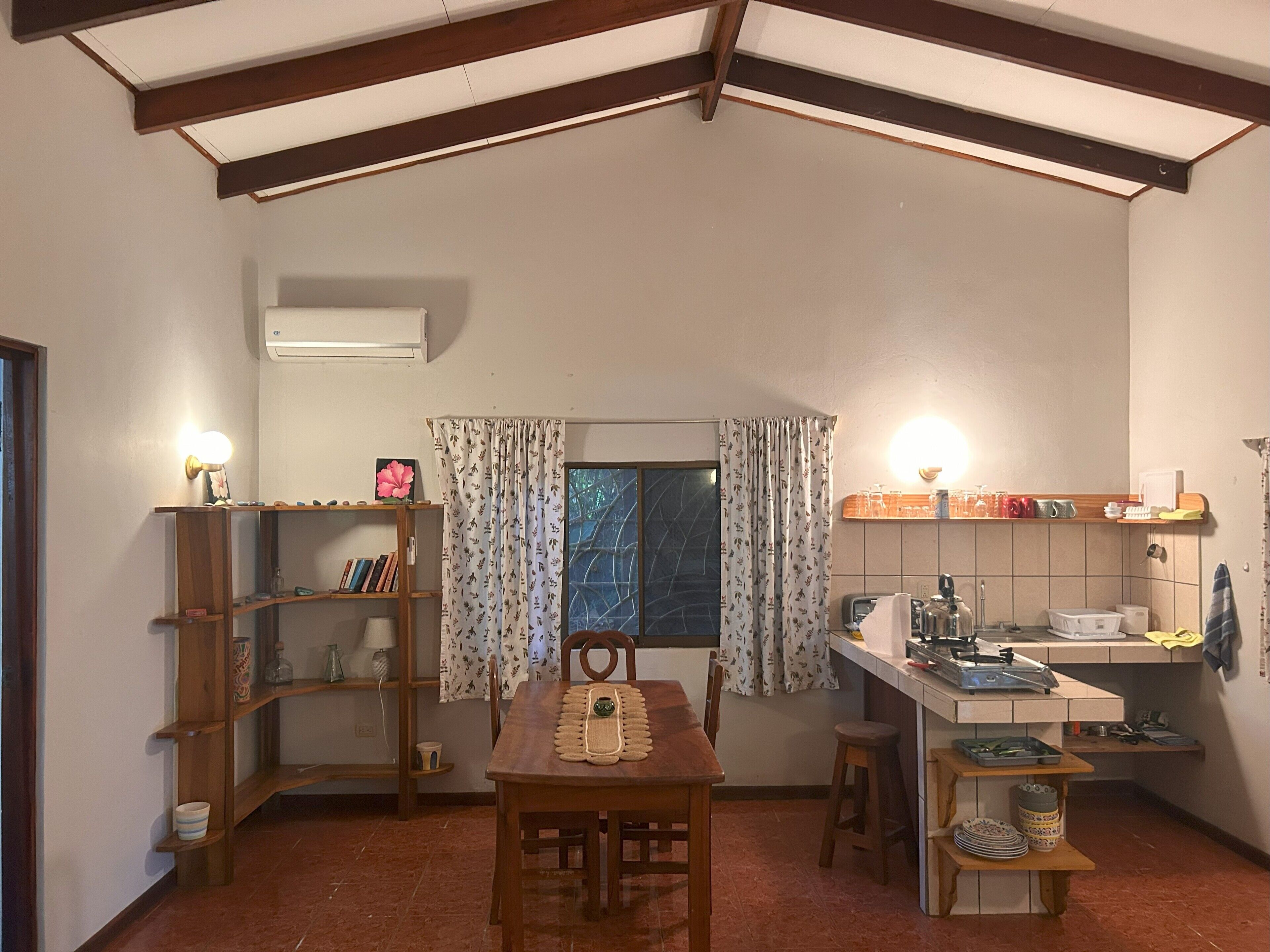 A compact kitchen with a dining table, wooden shelves, and a window with curtains.