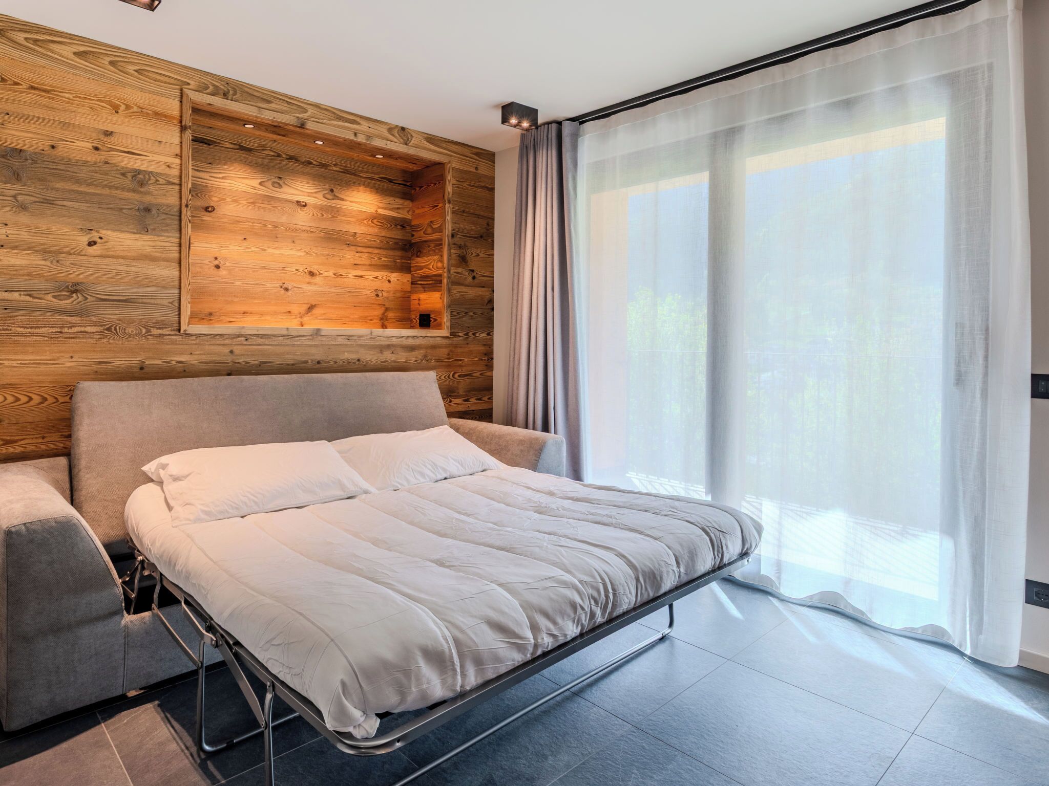 Une chambre à coucher moderne avec un lit, un banc intégré et une grande fenêtre ornée de rideaux légers.