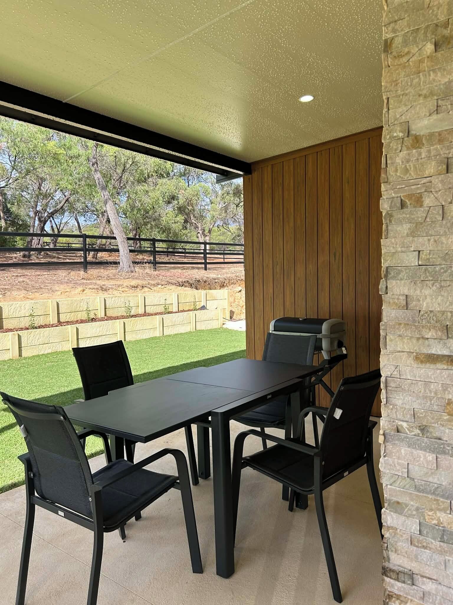 Une terrasse extérieure avec une table et des chaises, un barbecue et une vue sur une cour clôturée bordée d’arbres.