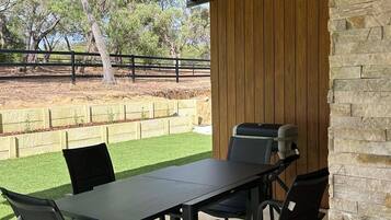 Une terrasse extérieure avec une table et des chaises, un barbecue et une vue sur une cour clôturée bordée d’arbres.