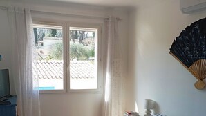 Un dormitorio con cama, ventana con cortinas, mesita de noche con lámpara y ventilador de pared.