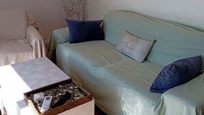 Una habitación con un sofá verde claro, dos almohadas azules, un adorno en la pared, una lámpara de pie alta y un pequeño caja de madera con controles remotos.