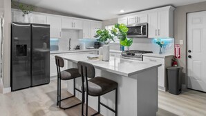 A modern kitchen with white cabinets, a stainless steel refrigerator, a central island with bar stools, and a microwave. - Grand North LV Escape w/ Pool, Spa, Arcade & Games! (North Las Vegas)