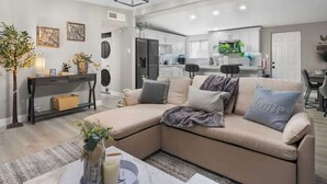 A modern living room with a beige sectional sofa, a coffee table with a vase, and a kitchen in the background. - Grand North LV Escape w/ Pool, Spa, Arcade & Games! (North Las Vegas)