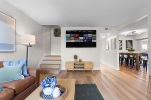 A modern living room with a flat-screen TV, a brown leather sofa, a wooden coffee table, and a dining area with a wooden table and chairs. - Grand Luxe Escape w/ 6 BR, Pool, Hot Tub, & Games! (North Las Vegas)