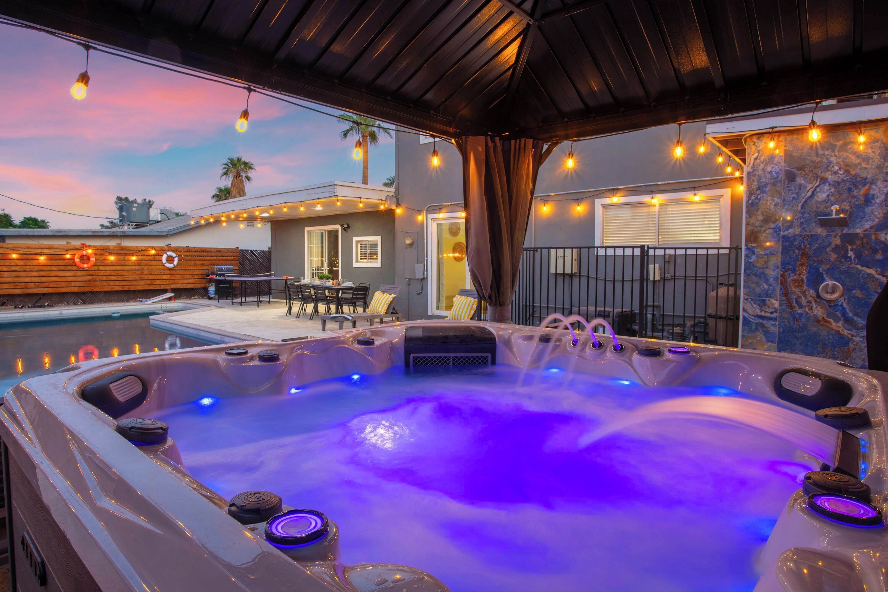 A hot tub with a waterfall feature, surrounded by a covered patio with string lights, a pool, and outdoor furniture.