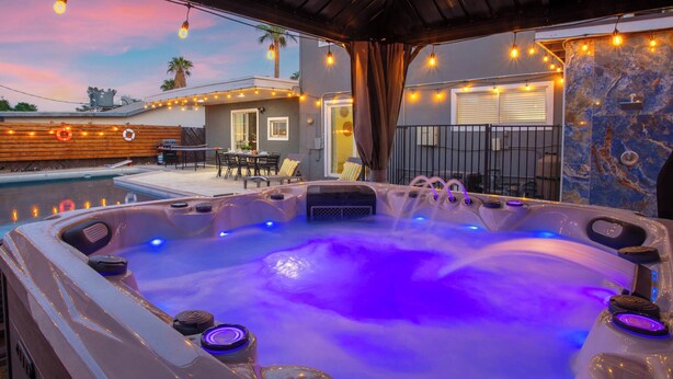 Un bain à remous agrémenté d’une cascade, entouré d’une terrasse couverte illuminée de guirlandes lumineuses, d’une piscine et de mobilier d’extérieur.