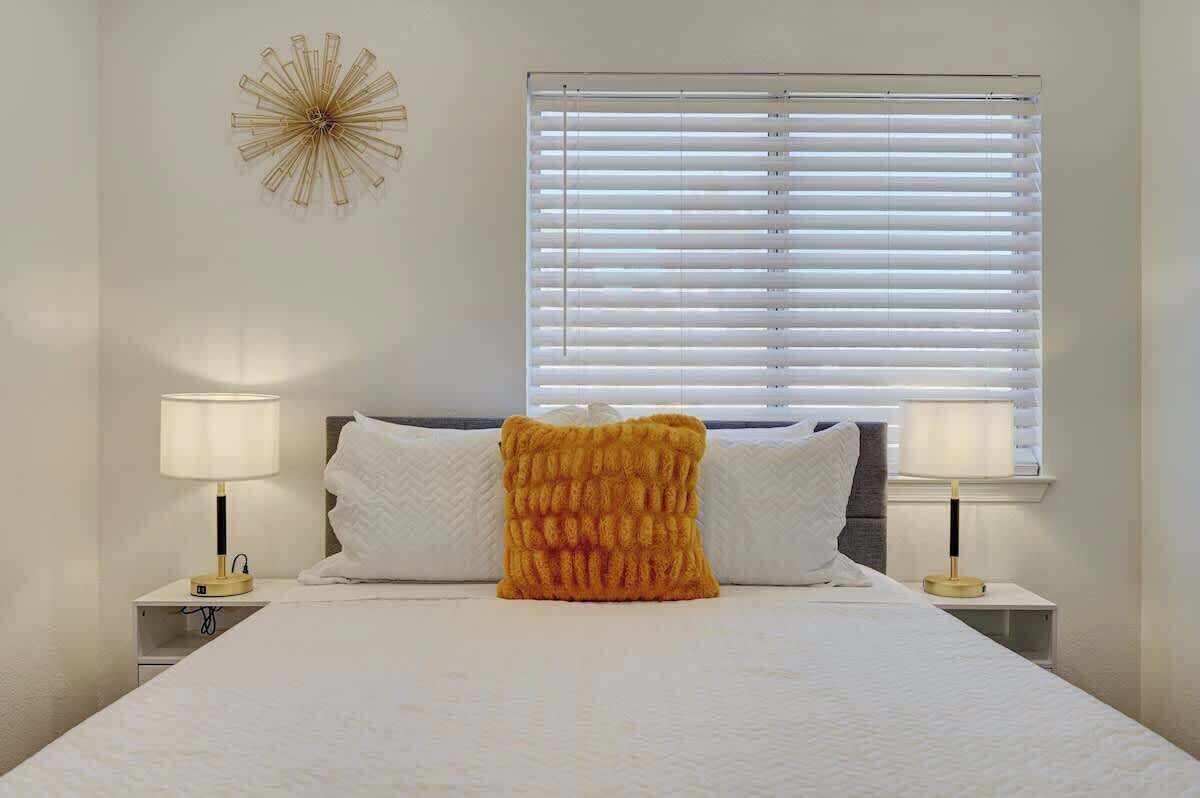 Ein ordentlich bezogenes Bett mit weißen Leinen, zwei Nachttischlampen, ein Fenster mit Jalousien und ein dekoratives Wandelement.
