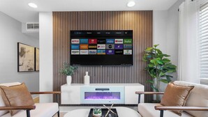 Een moderne woonkamer met een flatscreen televisie bevestigd aan een houten paneel, een haard onder de televisie, en twee witte banken met bruine kussens.