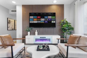 A modern living room with a flat-screen TV mounted on a wooden panel, a fireplace below, and two white sofas with brown cushions. - Modern 5BR Haven w/ Pool, Hot Tub, & Pool Table! (North Las Vegas)