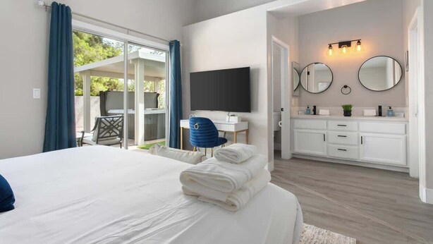 Ein Hotelzimmer mit Bett, Schreibtisch, Fernseher und einem Badezimmer mit Waschbecken und Spiegeln, sowie einer Glasschiebetür zu einem Außenbereich.