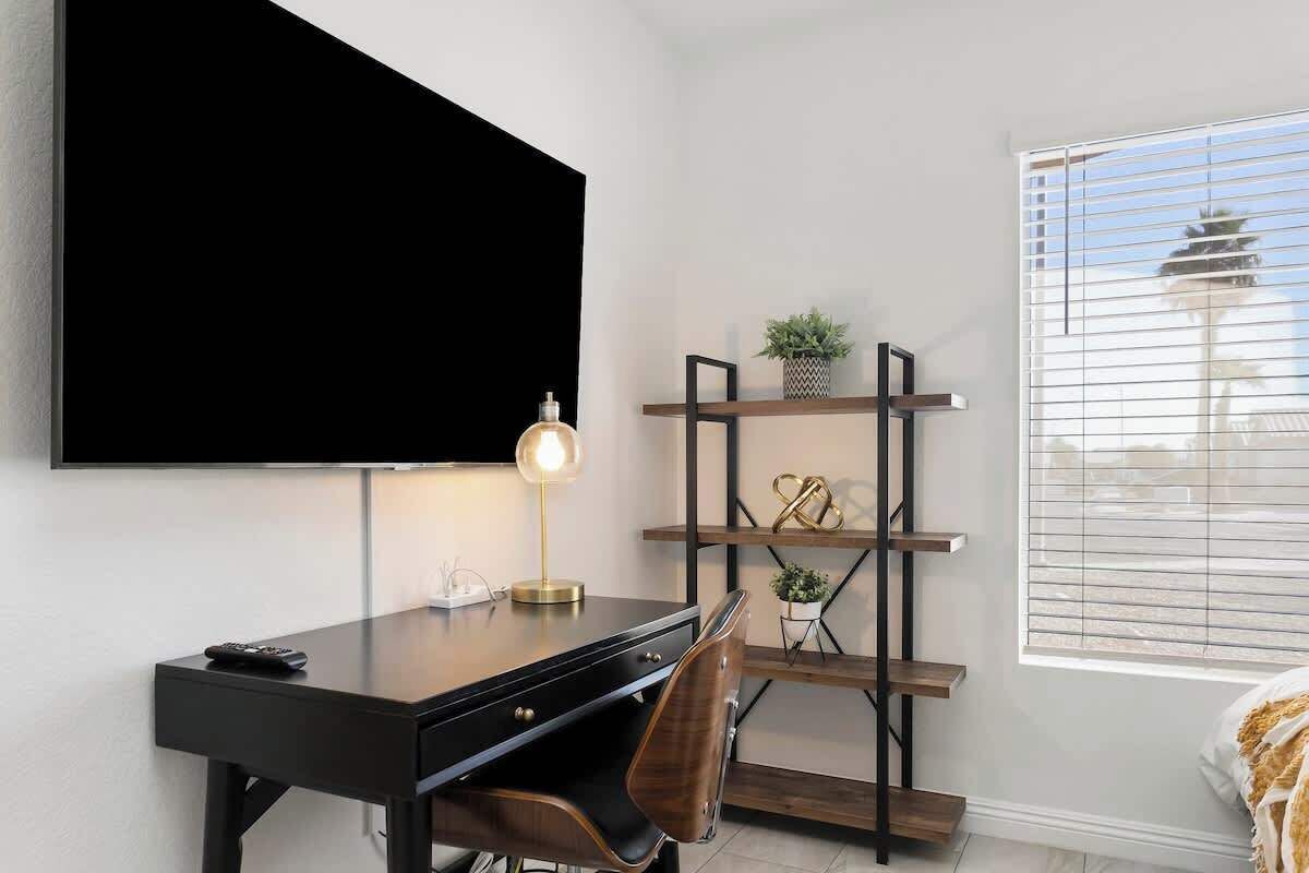 Une chambre moderne dotée d’une grande télévision, d’un bureau, d’une chaise, d’une table de chevet, d’une étagère avec des objets décoratifs et d’une fenêtre avec des stores.