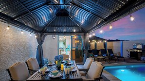 An outdoor patio with a pool, dining table, chairs, and a grill area.