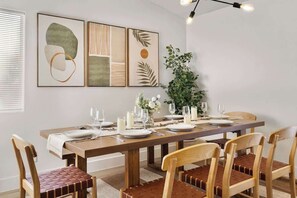 Ein Essbereich mit einem Holztisch, gedeckt für sechs Personen, umgeben von Holzsesseln mit braunen Kissen. Über dem Tisch hängen drei gerahmte Kunstwerke, rechts steht ein Topfpflanze und darüber eine Pendelleuchte.