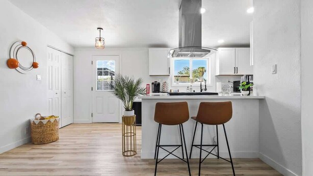 A modern kitchen with a central island, two bar stools, a hanging light fixture, and a view of the outdoors. - Radiant Pool Retreat: 4BR NLV Getaway (North Las Vegas)