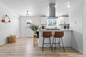 A modern kitchen with a central island, two bar stools, a hanging light fixture, and a view of the outdoors. - Radiant Pool Retreat: 4BR NLV Getaway (North Las Vegas)