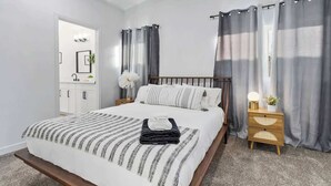 A bedroom with a bed, a bedside table with a lamp, a nightstand, a mirror, and a bathroom with a sink and cabinets. - Radiant Pool Retreat: 4BR NLV Getaway (North Las Vegas)