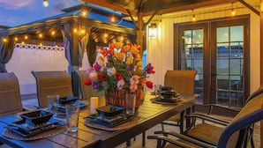 Un área de comedor al aire libre con una mesa dispuesta para dos, rodeada de sillas, un florero y luces colgantes.