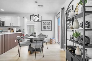 A modern kitchen with white cabinets, a dining area with a round table and chairs, and a black shelving unit with decorative items. - Chill&Play in 6BR Retreat w Pool, Hot Tub & Games! (North Las Vegas)