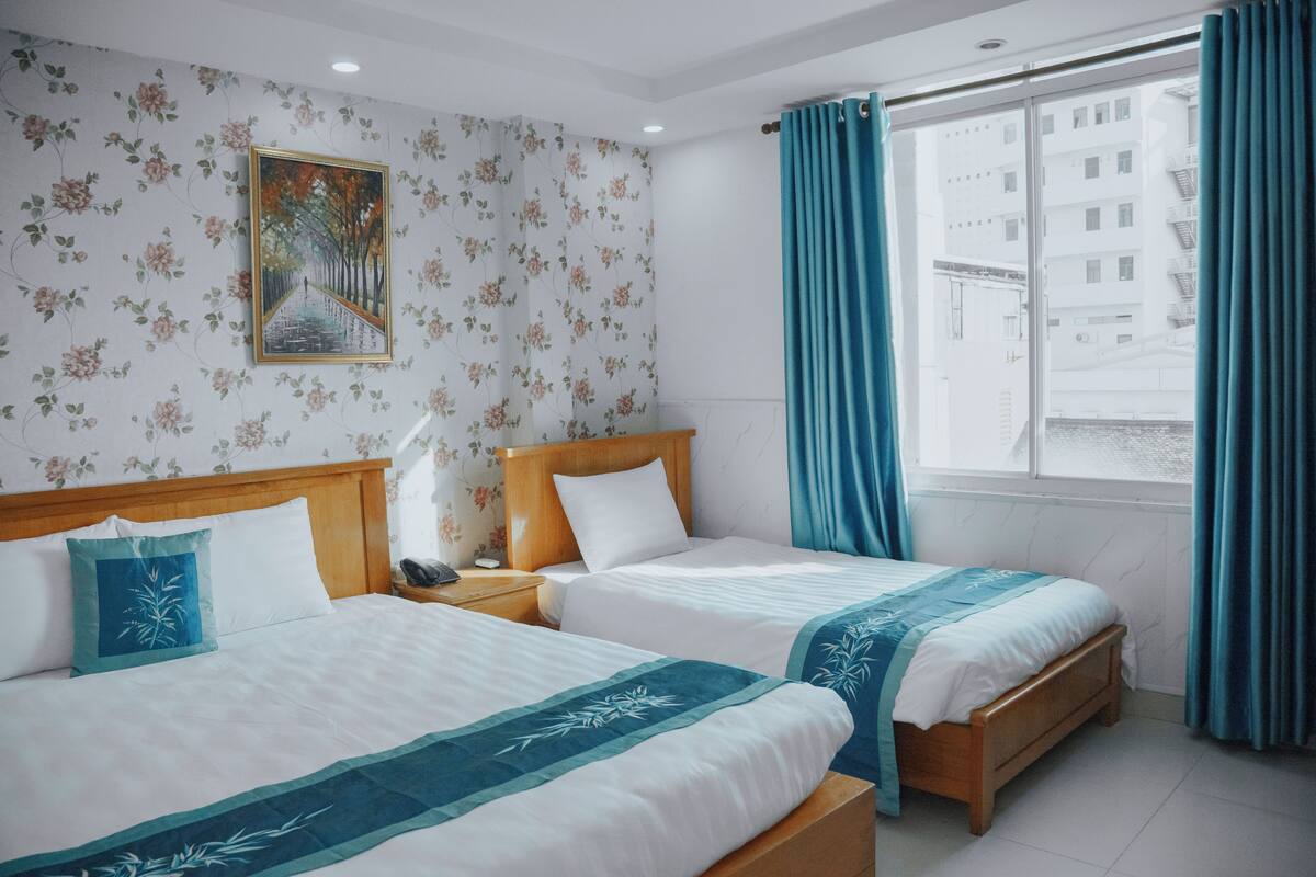Habitación de hotel con dos camas, cabecera de madera, mesita de noche, un cuadro en la pared y una ventana con cortinas.