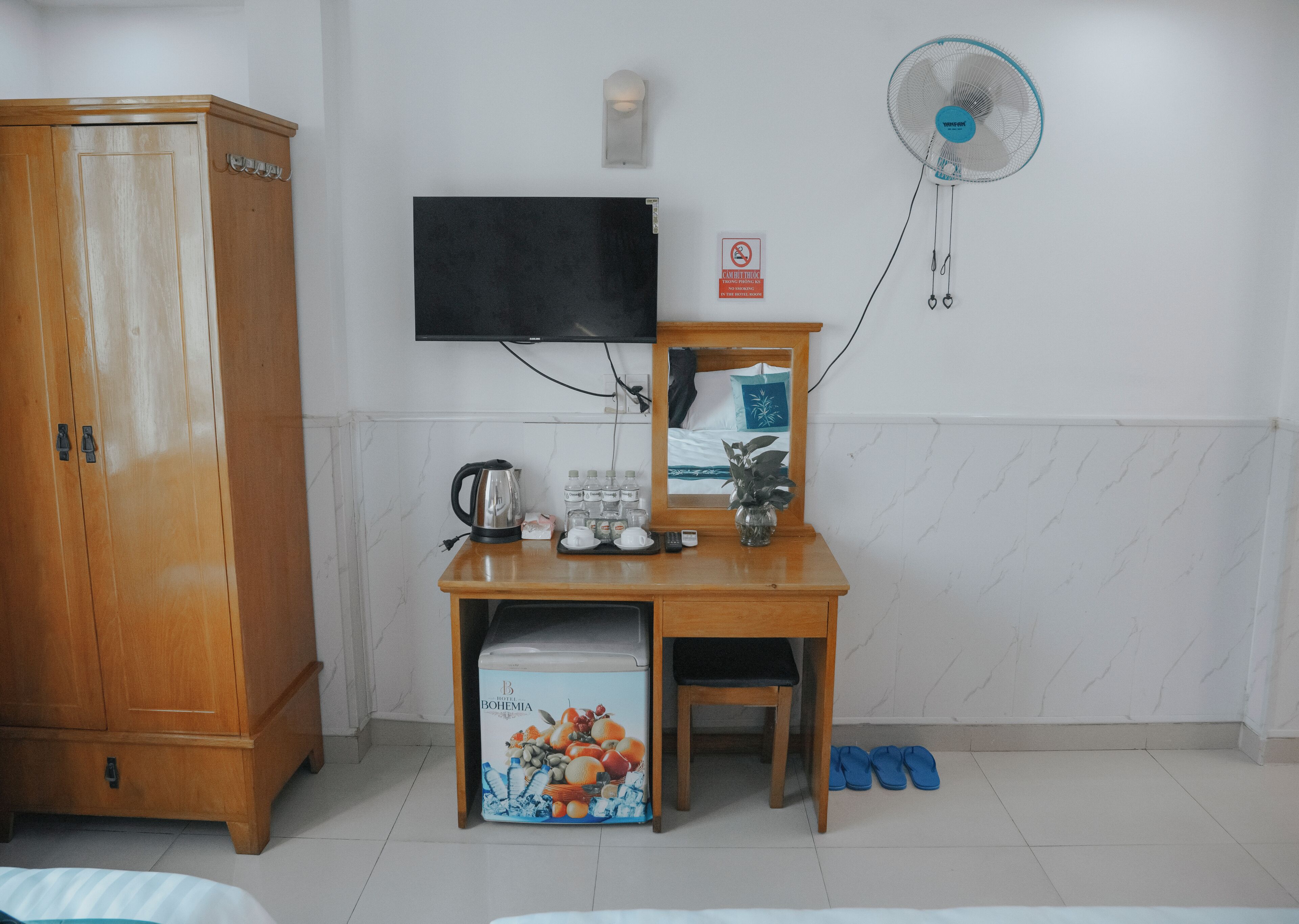 Una habitación con un escritorio de madera, televisor, espejo, armario, ventilador, un refrigerador pequeño y un par de pantuflas.