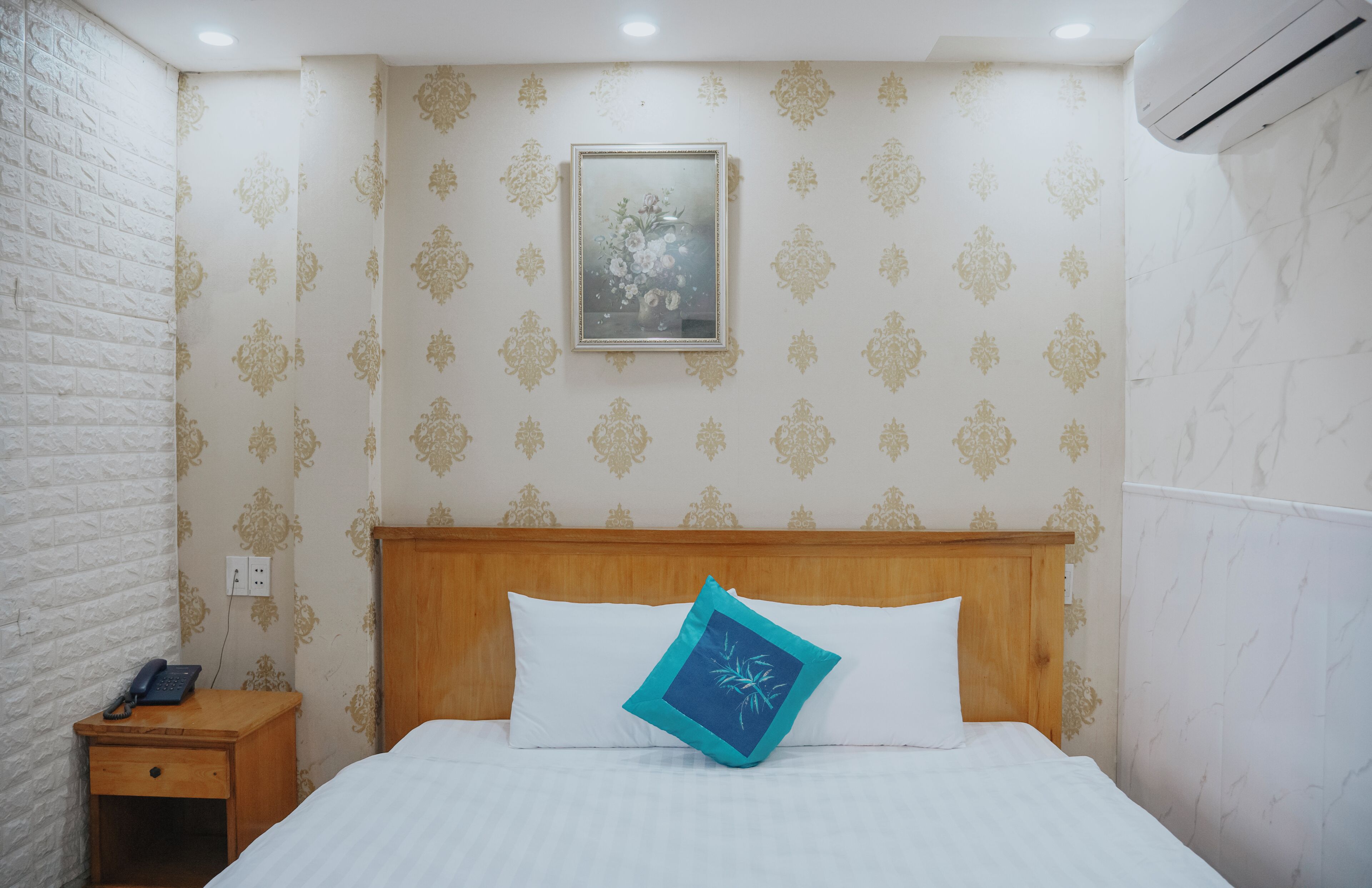 A hotel room with a wooden headboard, a bed with white bedding, a nightstand with a phone, and patterned wallpaper.