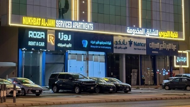 Un edificio con varios letreros, incluyendo 'Nukhabat Al-Jawiy Serviced Apartments' y servicios de 'Rent a Car'.