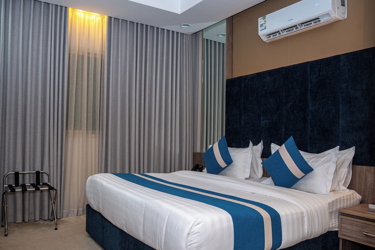 Ein Hotelzimmer mit einem großen Bett, einem Nachttisch, einem Sessel, einer Wand-Klimaanlage und einem Fenster mit Vorhängen.