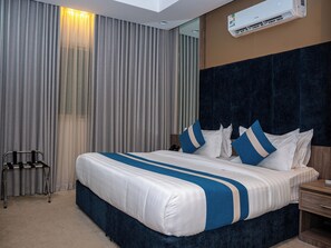 Ein Hotelzimmer mit einem großen Bett, einem Nachttisch, einem Sessel, einer Wand-Klimaanlage und einem Fenster mit Vorhängen.