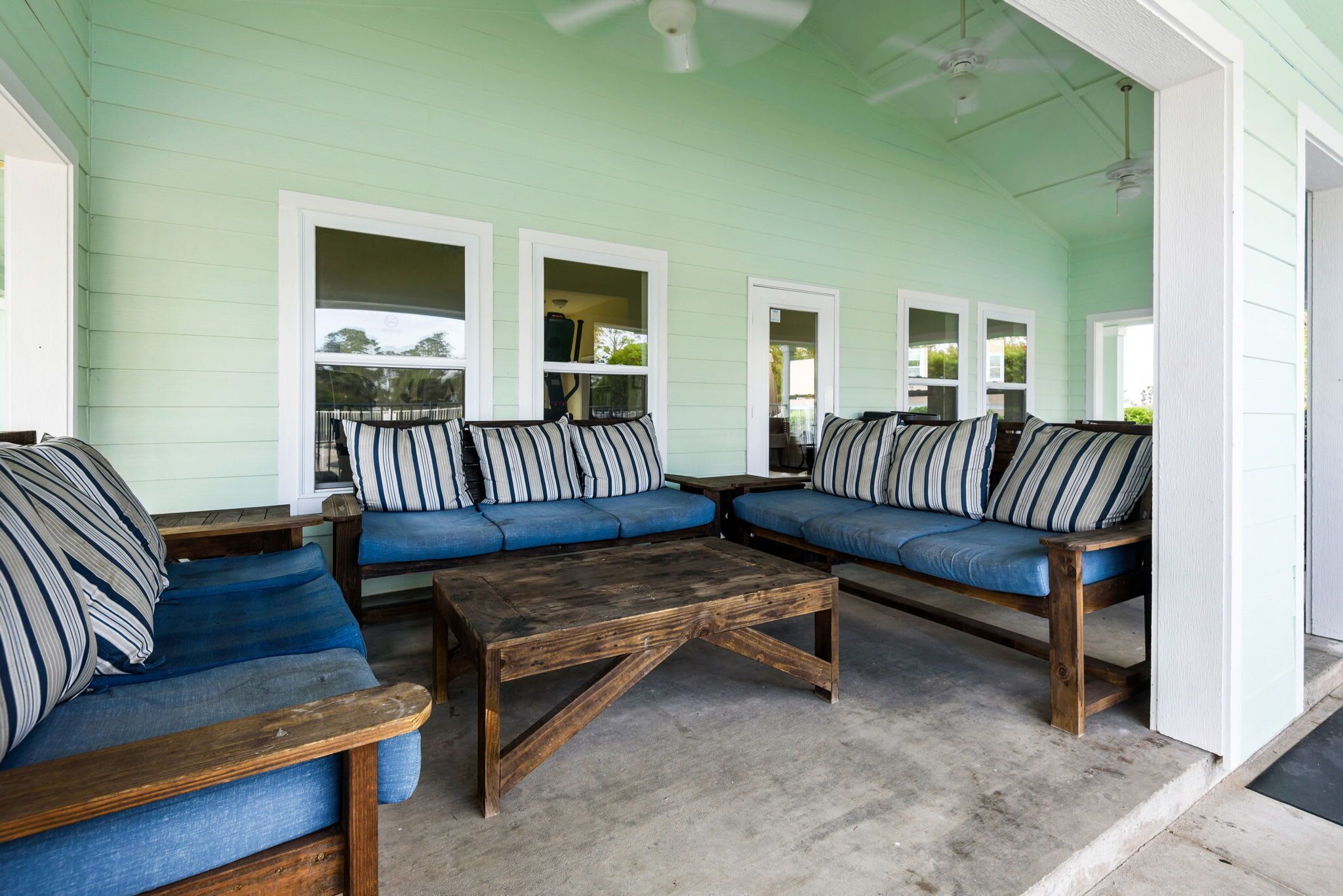 A covered porch with blue cushioned seating and striped pillows, wooden tables, and white-framed windows.