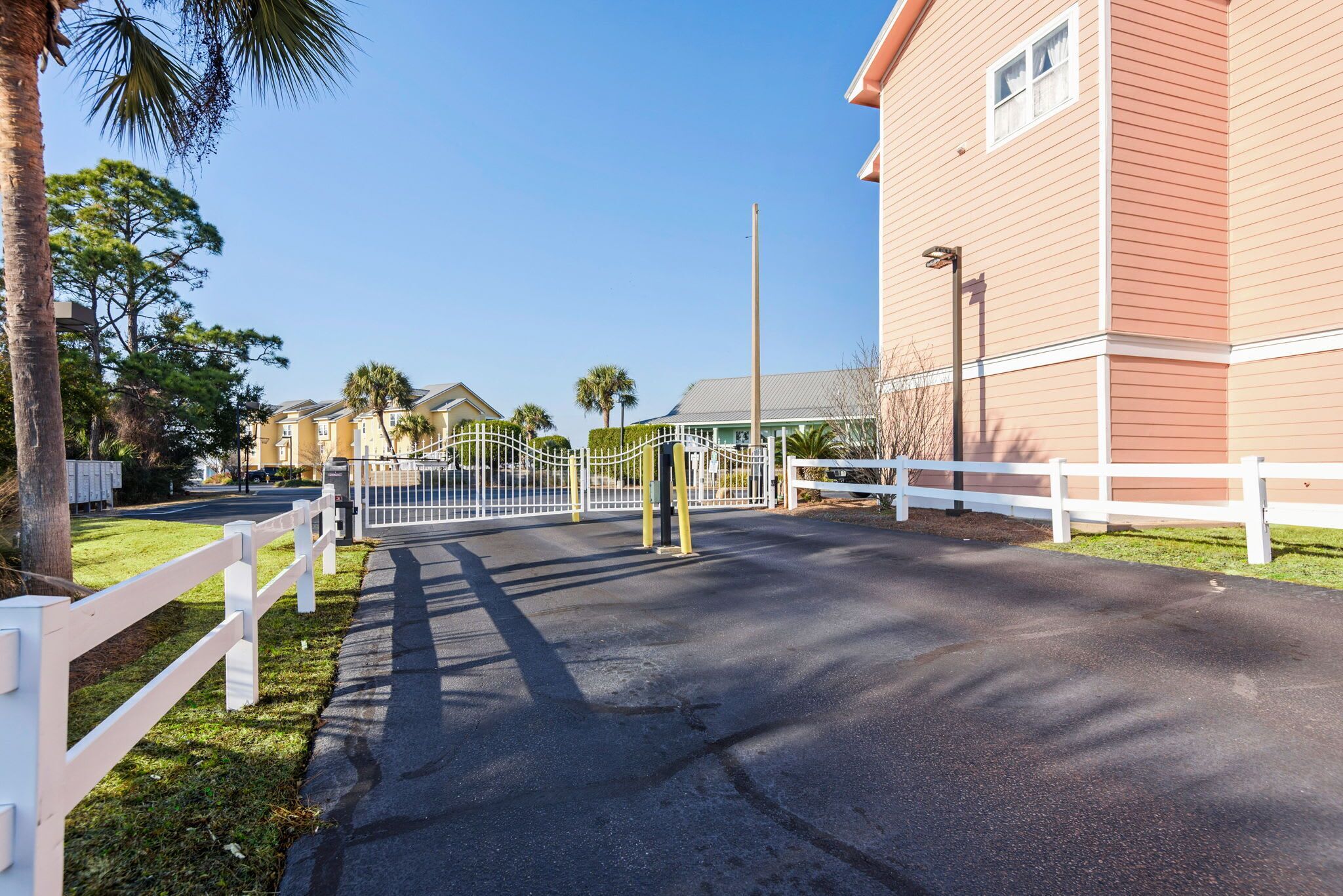 A gated entrance to a residential area with a paved road, white fences, and houses with balconies.