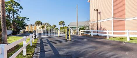A gated entrance to a residential area with a paved road, white fences, and houses with balconies.