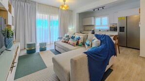 A modern living room with a sofa, ottomans, a TV, and a kitchen in the background. - Tracadero Lodge PH C7 - 2 Hab Apto (Dominicus)