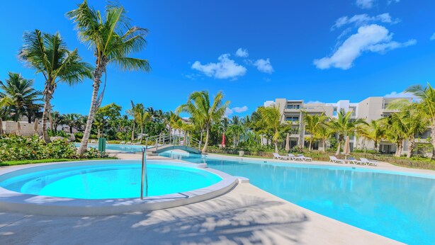 A swimming pool with a circular section, surrounded by palm trees and lounge chairs, with a clear blue sky in the background. - Tracadero Lodge PH C7 - 2 Hab Apto (Dominicus)