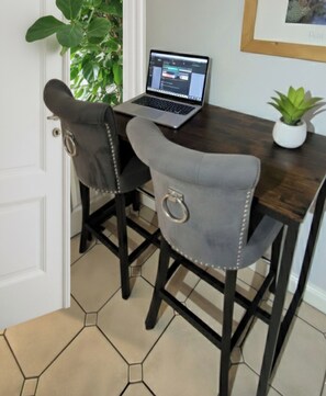 Un coin repas avec une table en bois foncé, deux chaises grises à revêtement en tissu et ornées d’anneaux en métal, un ordinateur portable posé sur la table et une plante en pot sur la droite.