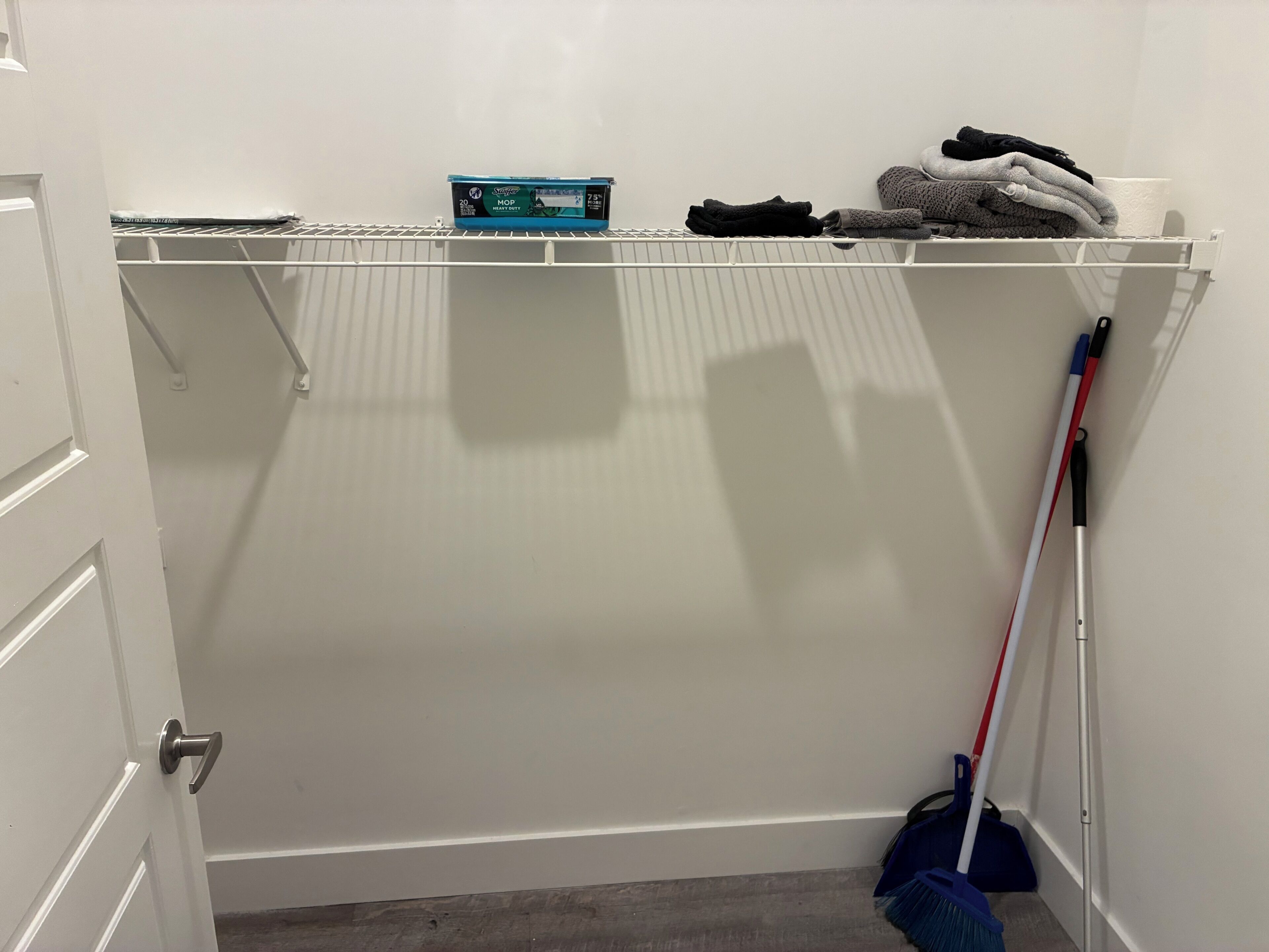 Ein Schrank mit einem Einlegeboden, der eine Kiste, gefaltete Kleidung und einen Besen beherbergt.