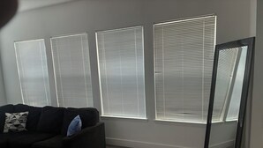Una habitación con un sofá negro, tres ventanas con persianas y un espejo.