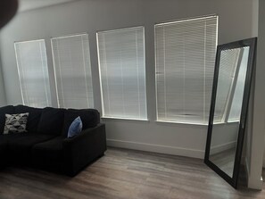 A room with a black sofa, three windows with blinds, and a mirror. - Live Steps from the Action: Stylish 2BR/2BA in Downtown Atlanta for FIFA Fans! (Atlanta)