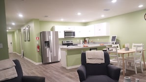 A modern kitchen with white cabinets, a stainless steel refrigerator, a microwave, and a stove. There is a dining area with a table and chairs, and a dark armchair. - Unwind in this serene thoughtfully designed home nestled in a quiet neighborhood (Poinciana)