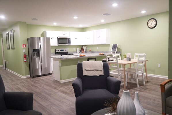 A modern kitchen with stainless steel appliances, a dining area with a wooden table and chairs, and a living room with a dark armchair and decorative vases.