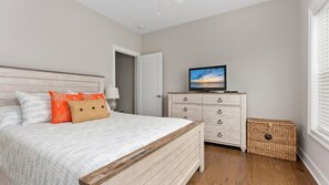 Ein Schlafzimmer mit einem Bett, einem Fernseher auf einer Kommode, einem Korbwäscheschrank und einer Tür zu einem anderen Raum.