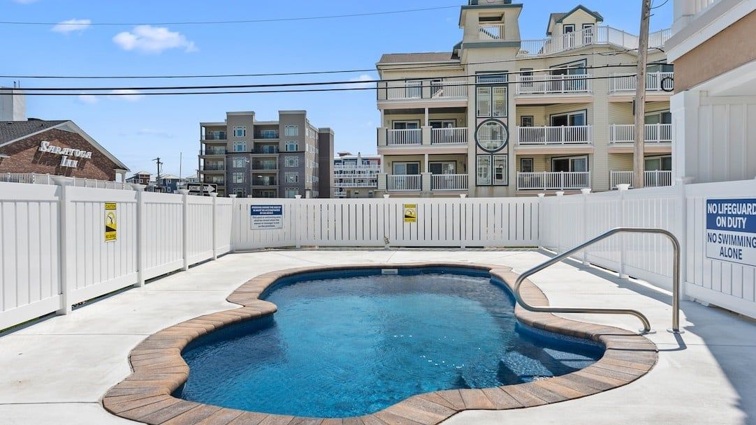 Una piscina con una cerca blanca, un cartel de salvavidas y un edificio con balcones al fondo.
